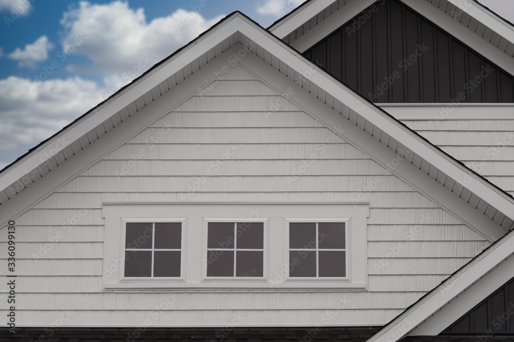 Closeup triple gable facade with thick white frame triple attic window, white horizontal vinyl siding, dark vertical siding new construction East Coast American single family home with cloudy blue sky