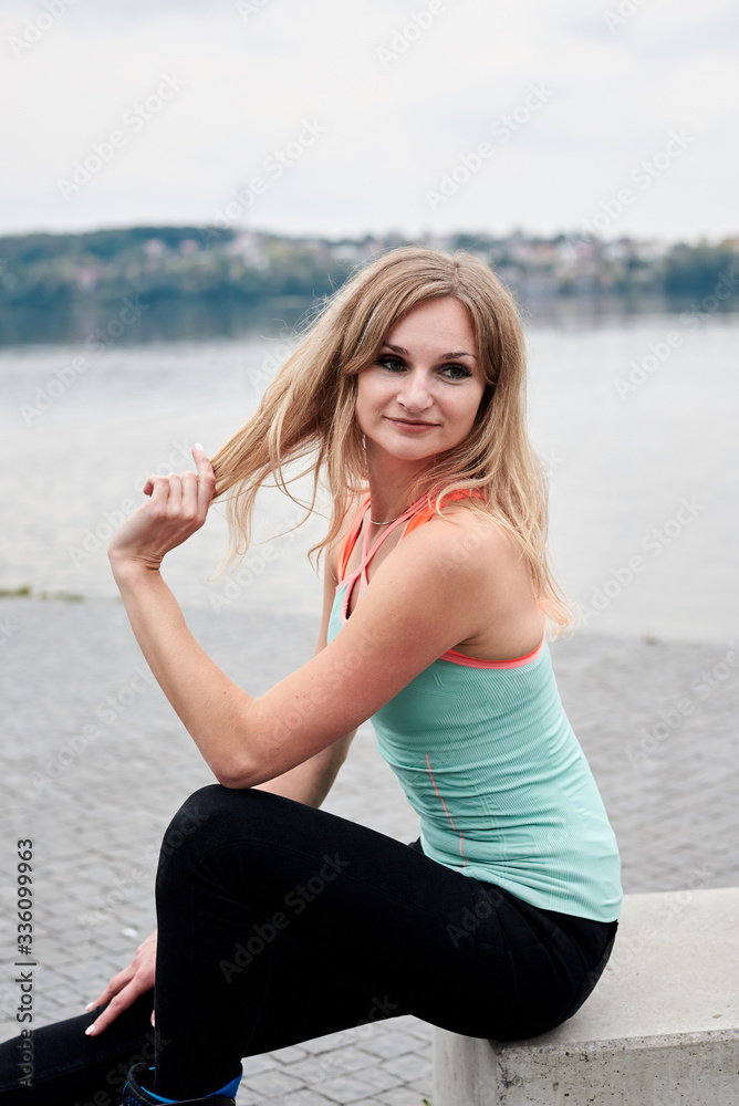 Young blond fit slim woman, sitting in front of city lake in summer. Three-quarter picture of fitness trainer, wearing black leggings and mint top, posing. Healthy lifestyle concept, wellness.