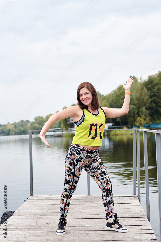Young brunette sportswoman, wearing yellow top with zumba sign and colorful  leggings on wooden pier. Full-length portrait of Zumba fitness instructor,  dancing by city lake. Healthy lifestyle concept. Stock Photo