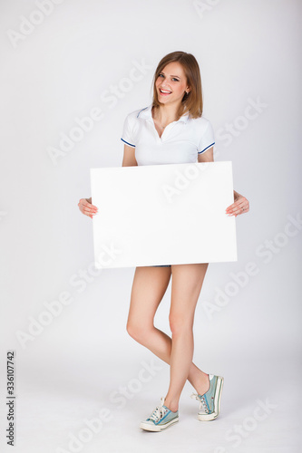 Beautiful young gir in denim shorts and a T-shirth on a white background holding big white blank card. Pretty student girl having fun and holding white empty signboard background with copy space.