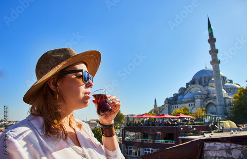 Photography Happy attractive woman tourist in hat, drinks traditional turkish tea (armudas) against the background of Suleymaniye mosque, Istanbul, Turkey