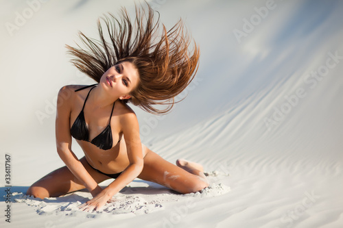 Obraz na płótnie Attractive girl in black swimsuit posing kneeling in sand, waving her hair and looking in camera. Photo outdoors w salonie