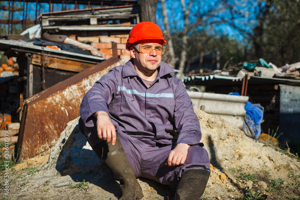A young construction worker in an orange helmet and protective glasses sits on a pile of sand. Building. Portrait.