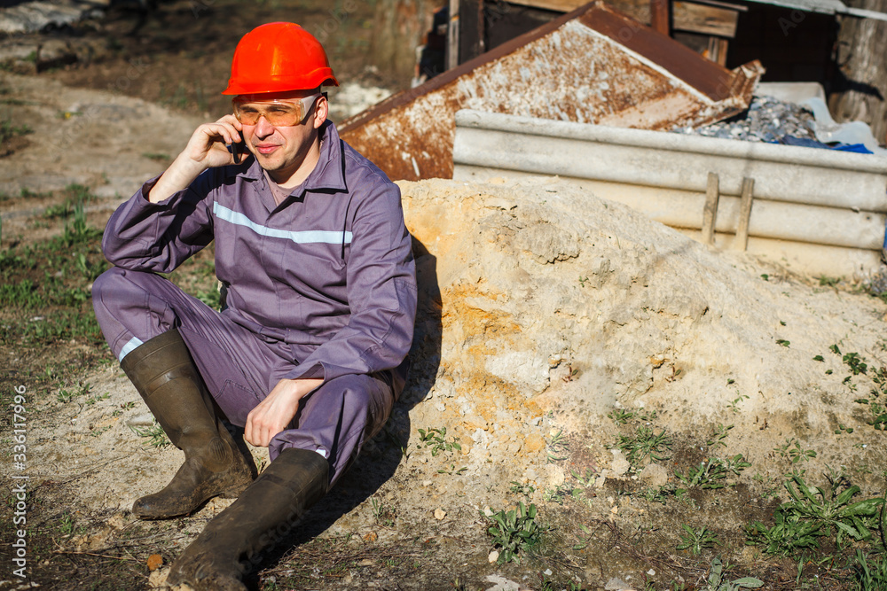 A young construction worker in an orange helmet and protective glasses sits on a pile of sand. Building. Portrait. The builder is talking on the phone. A young construction worker in an orange helmet and protective glasses sits on a pile of sand. Building. Portrait. The builder is talking on the phone.