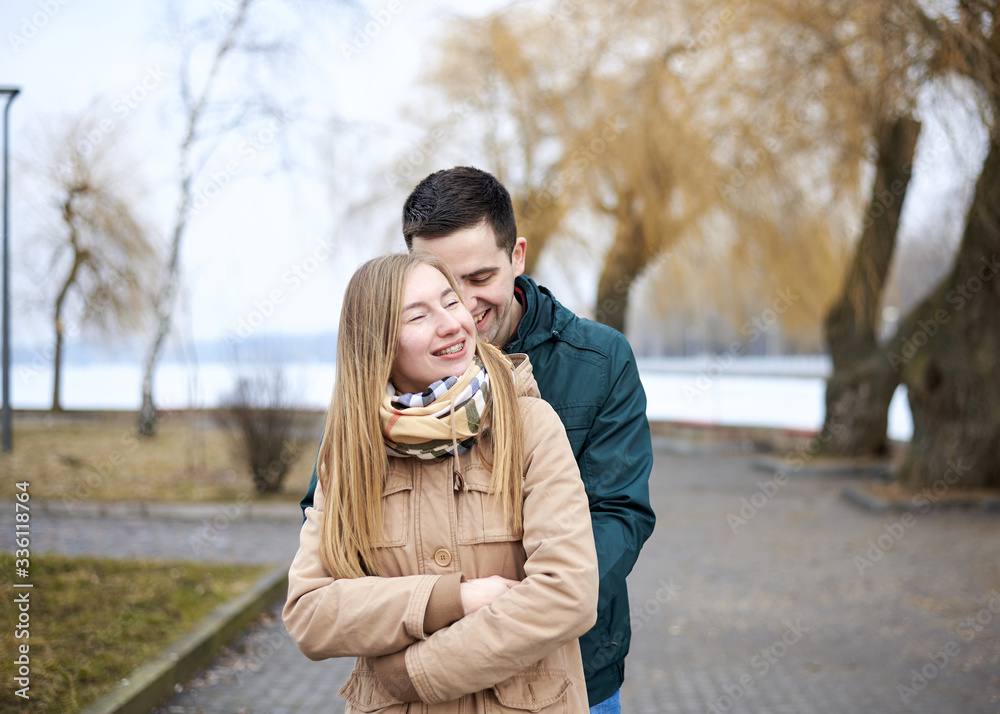 Young brunette man, wearing green jacket, standing behind young blond woman, wearing beige jacket, hugging her, smiling, kissing. Romantic Valentines day celebration Young brunette man, wearing green jacket, standing behind young blond woman, wearing beige jacket, hugging her, smiling, kissing. Romantic Valentines day celebration