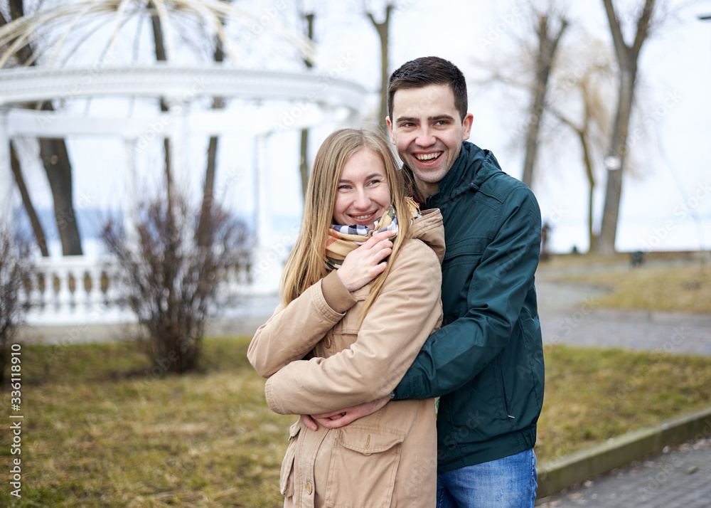 Young brunette man, wearing green jacket, standing behind young blond woman, wearing beige jacket, hugging her, smiling, kissing. Romantic Valentines day celebration Young brunette man, wearing green jacket, standing behind young blond woman, wearing beige jacket, hugging her, smiling, kissing. Romantic Valentines day celebration