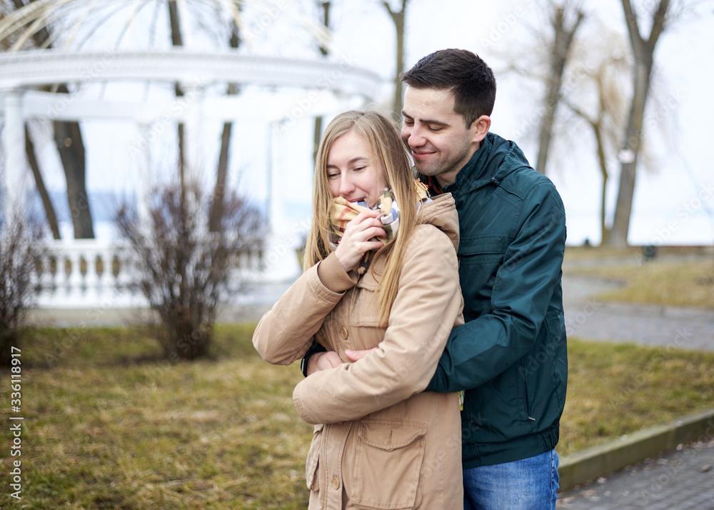 Fototapeta premium Young brunette man, wearing green jacket, standing behind young blond woman, wearing beige jacket, hugging her, smiling, kissing. Romantic Valentines day celebration
