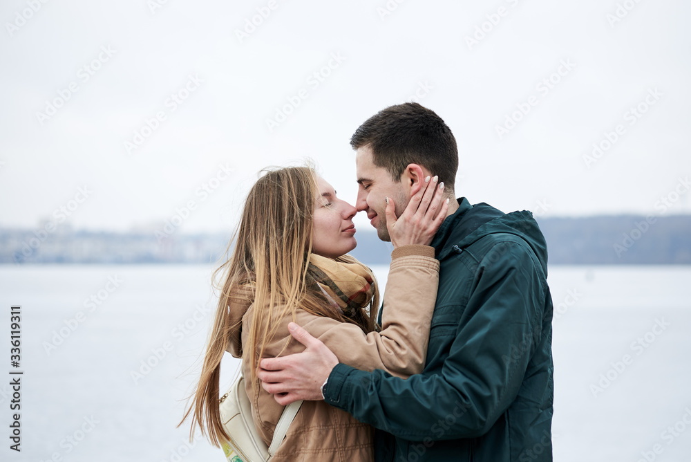 Young couple in love, wearing beige and green casual jackets, standing in front of frozen lake hugging, kissing each other in winter. Valentines day romantic celebration outside Young couple in love, wearing beige and green casual jackets, standing in front of frozen lake hugging, kissing each other in winter. Valentines day romantic celebration outside