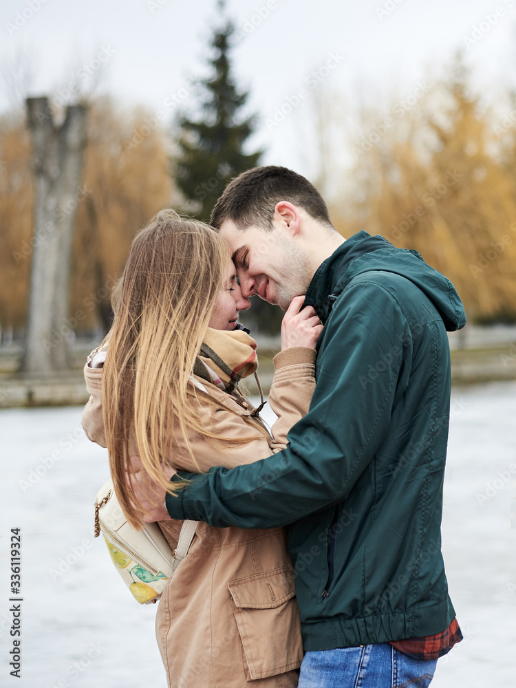 Fototapeta premium Young couple in love, wearing beige and green casual jackets, standing in front of frozen lake hugging, kissing each other in winter. Valentines day romantic celebration outside