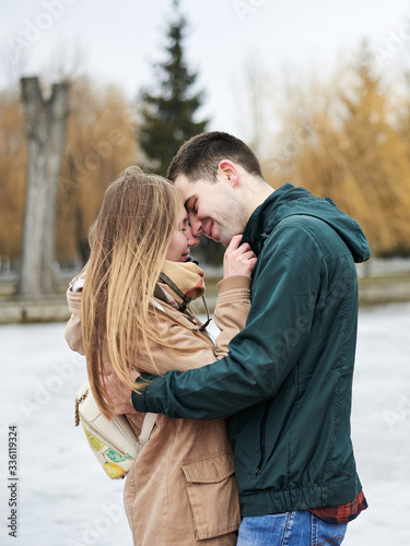 Young couple in love, wearing beige and green casual jackets, standing in front of frozen lake hugging, kissing each other in winter. Valentines day romantic celebration outside © Natalia