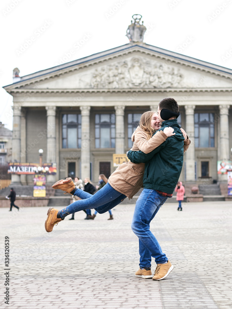 Young couple in love is having fun in the city center in front of old grey building. Brunette man is holding and spinning blond woman. Romantic Valentines day celebration