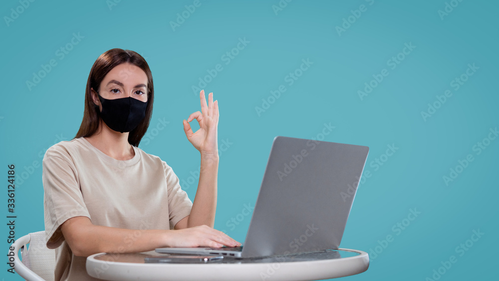 Pretty brunette female works with her computer isolated, she shows ok sign and wear medical mask, isolated over blue bg
