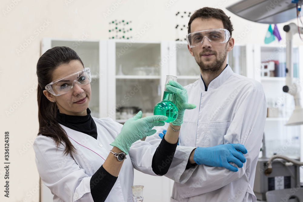 Obraz premium Two chemists work in a modern chemical laboratory. Senior chemist and laboratory assistant study the green solution in the flask.