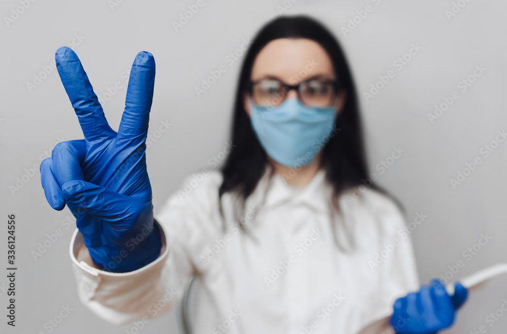 Young doctor in a white coat, blue protective medical mask and purple rubber gloves shows victory gesture. Girl nurse. The concept of the end of quarantine, the pandemic of the coronavirus COVID-19. Young doctor in a white coat, blue protective medical mask and purple rubber gloves shows victory gesture. Girl nurse. The concept of the end of quarantine, the pandemic of the coronavirus COVID-19.