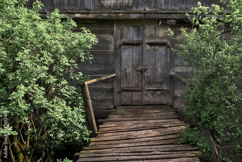 Old wooden grey hut in the village. Old time watermill among green trees in coutryside. Doors of ancient wooden building 