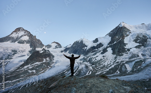 Happy traveller welcoming night in the mountains, standing with his back to the camera with open hands looking at glacier and high snowy peaks of Pennine Alps © anatoliy_gleb