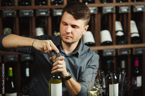 Portrait of young caucasian sommelier uncorked a bottle of white wine at the table on wine shelf background. photo