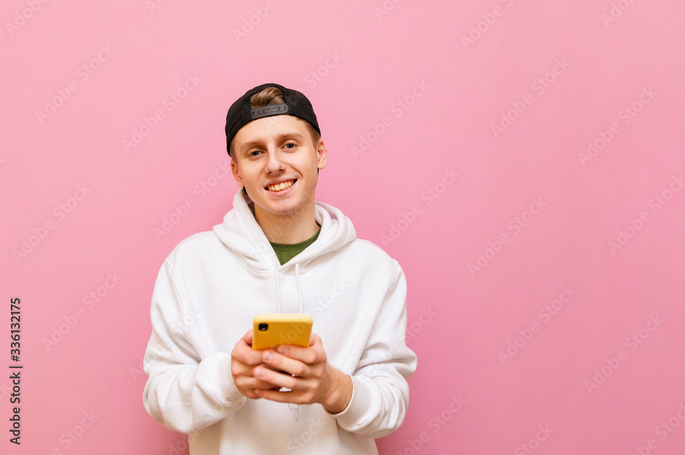 Smiling guy in a cap and white hoodie stands on a pink background with a smartphone in his hand and looks into the camera with a happy face. Cheerful student uses internet on smartphone. isolated Smiling guy in a cap and white hoodie stands on a pink background with a smartphone in his hand and looks into the camera with a happy face. Cheerful student uses internet on smartphone. isolated