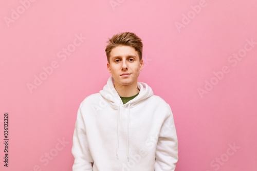 Portrait of attractive guy in casual clothes stands on pink background, looks into camera with serious face, wears white hoodie. Cute young man with blond hair posing, isolated on pink background.