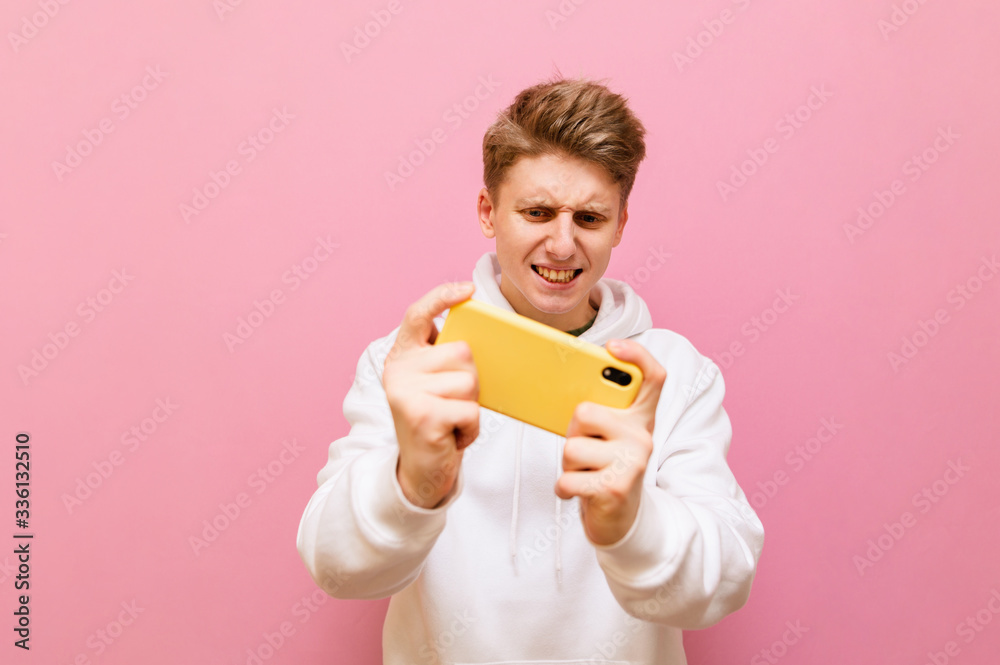 Positive young gamer in white hoodie playing mobile games with on smartphone on pink background. Concentrated teenager playing online games on a smartphone, isolated on a pink background. Copy space Positive young gamer in white hoodie playing mobile games with on smartphone on pink background. Concentrated teenager playing online games on a smartphone, isolated on a pink background. Copy space