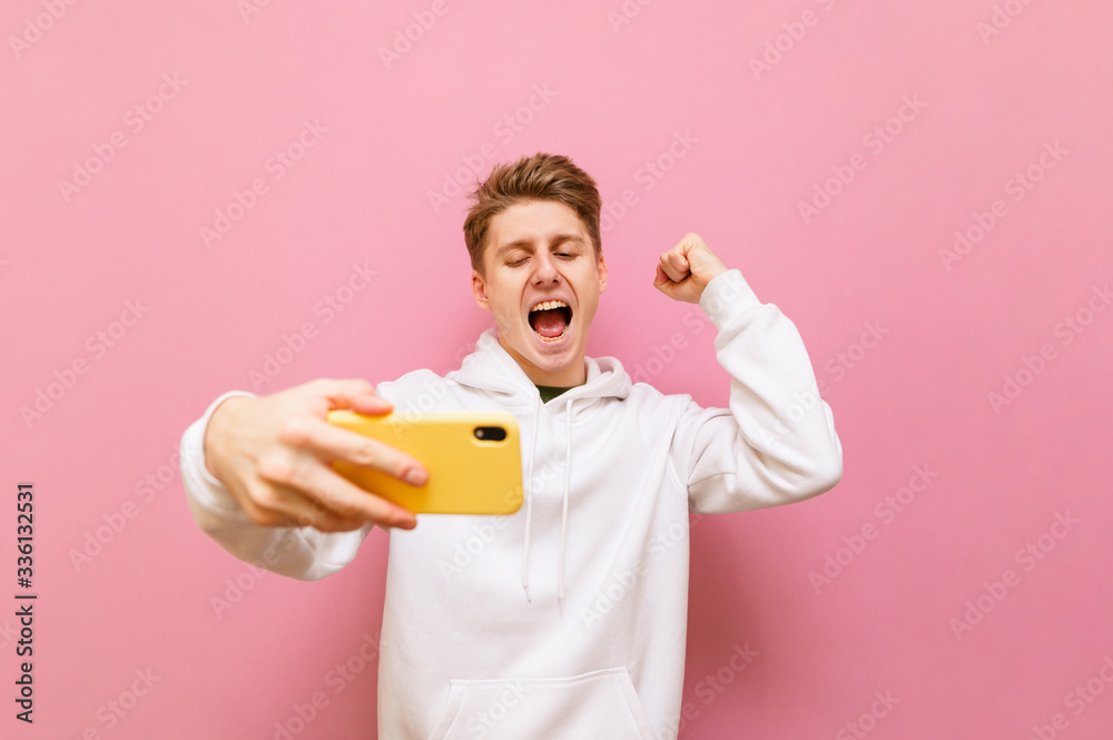Joyful guy gamer stands with a smartphone in his hand on a pink background and is emotionally pleased to win with hand raised and a smile on face.Happy teenager rejoices in winning the online game Joyful guy gamer stands with a smartphone in his hand on a pink background and is emotionally pleased to win with hand raised and a smile on face.Happy teenager rejoices in winning the online game