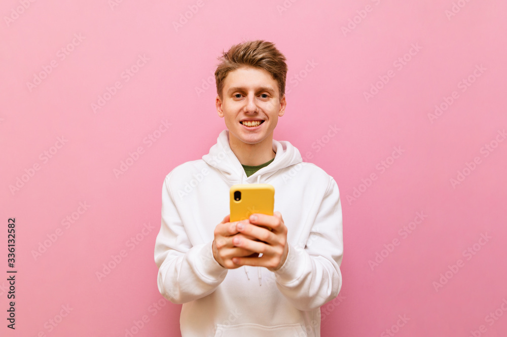 Cheerful blond guy in white casual clothes stands on a pink background with a smartphone in his hands, looks into the camera and smiles. Positive teenager using smartphone ,isolated. Copy space