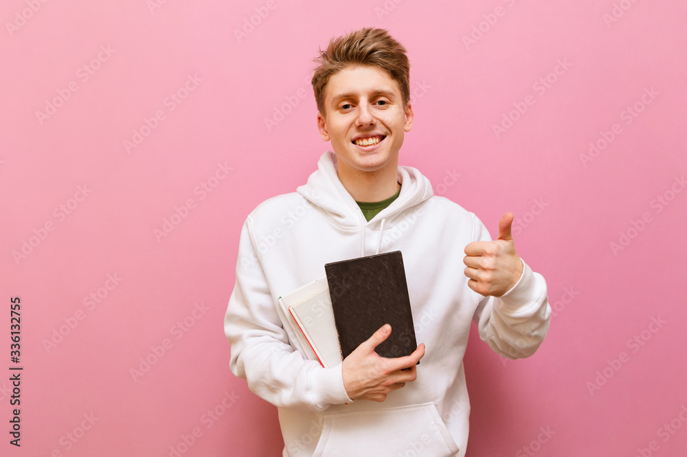 Happy student in white roe deer stands on a pink background with books in his hands, looks into the camera, smiles and shows his thumb up. Smiling teenager with books posing on camera, isolated.