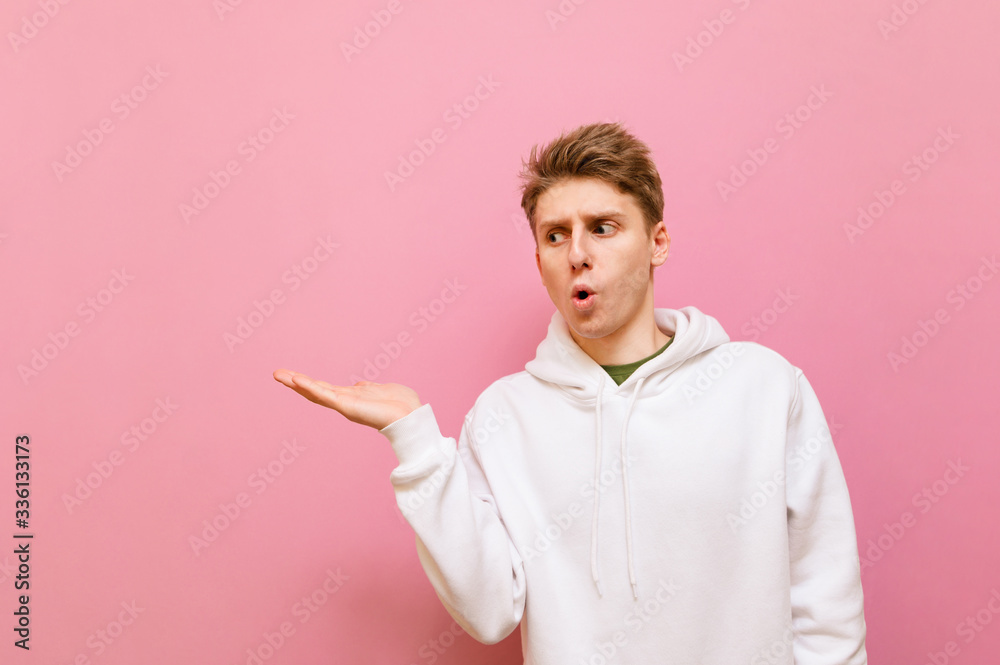 Funny guy stands on a pink background and looks to the side holding a copy space with a surprised face. Shocked young man holding an empty seat on his hand, Isolated. Funny guy stands on a pink background and looks to the side holding a copy space with a surprised face. Shocked young man holding an empty seat on his hand, Isolated.