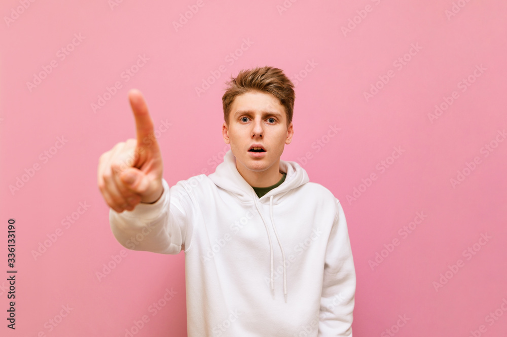 Surprised young man in white hoodie stands on pink background and touches finger to camera. Emotional guy in casual clothes pushes his finger forward and looks with shocked face. Copy space Surprised young man in white hoodie stands on pink background and touches finger to camera. Emotional guy in casual clothes pushes his finger forward and looks with shocked face. Copy space