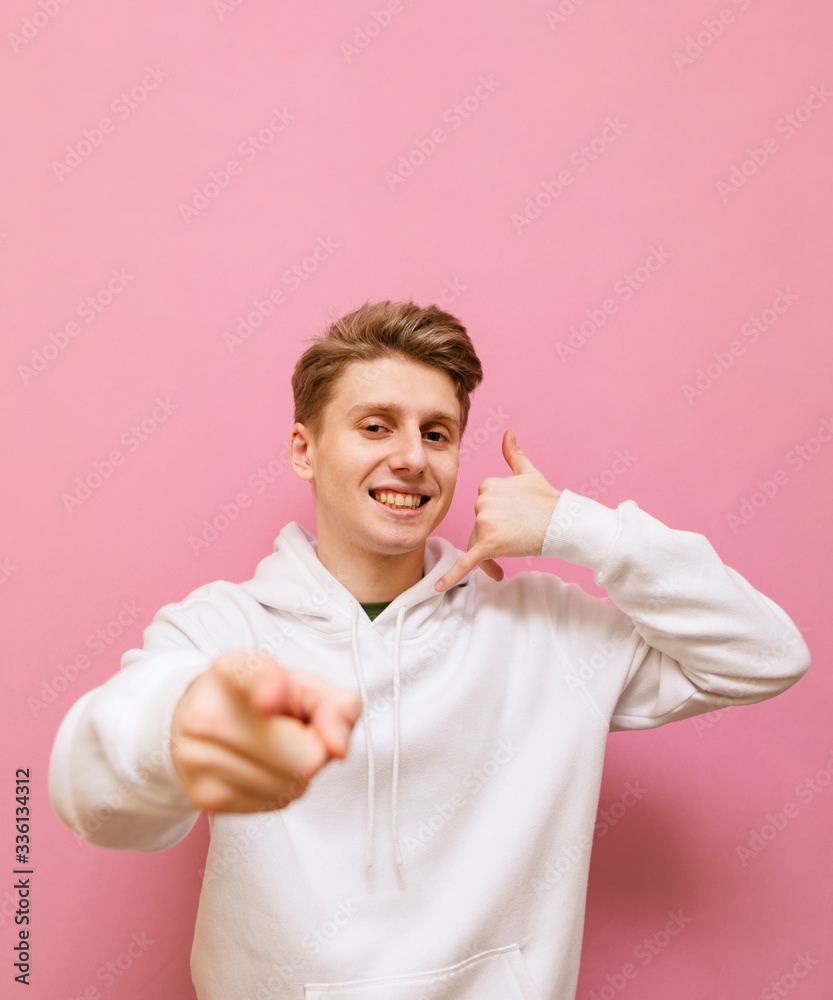 Smiling guy in white sweatshirt shows call me gesture on pink background, looks into camera with happy face. Cheerful guy shows a ringing gesture and a finger at you. Copy space Smiling guy in white sweatshirt shows call me gesture on pink background, looks into camera with happy face. Cheerful guy shows a ringing gesture and a finger at you. Copy space