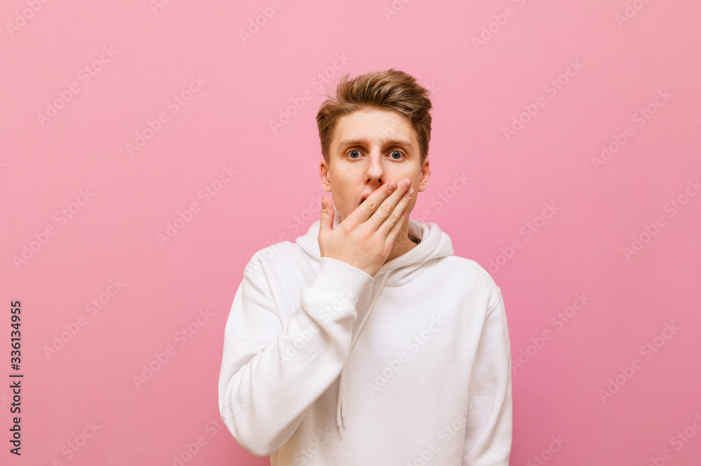 Astonished young man stands on a pink background and looks at the camera with a surprised face. Surprised guy student isolated on pink, covered his mouth from shock, copy space
