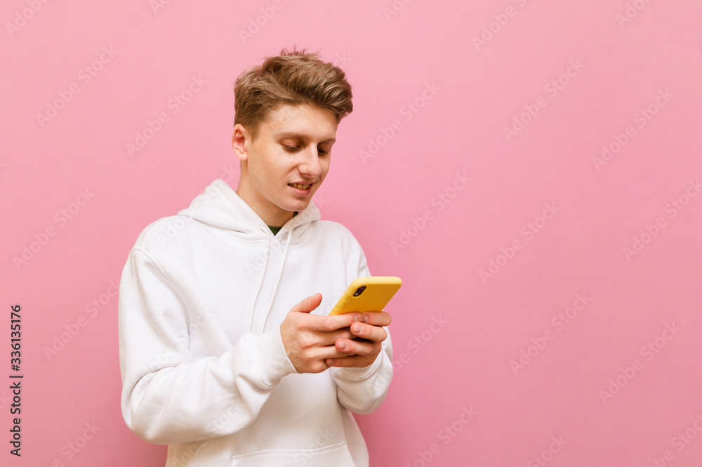 Cheerful guy in white hoodie is using smartphone with smile on face on pink background. Positive young man talking on the internet on a smartphone with a smile on his face. Copy space Cheerful guy in white hoodie is using smartphone with smile on face on pink background. Positive young man talking on the internet on a smartphone with a smile on his face. Copy space