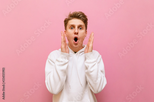 Shocked young man standing on a pink background, looking mad at the camera and putting his hands to his face. Portrait of surprised guy in white hoodie looking into camera with crazy face. Copy space