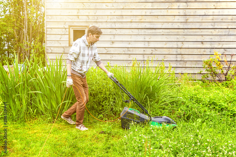 Man cutting green grass with lawn mower in backyard. Gardening country lifestyle background. Beautiful view on fresh green grass lawn in sunlight, garden landscape in spring or summer season