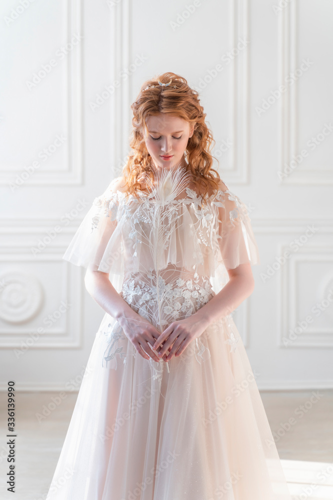 Beautiful natural redhead girl bride, with nude makeup, wearing a white dress, stands at the white wall, holding a white feather in her hands in light interior. Beautiful natural redhead girl bride, with nude makeup, wearing a white dress, stands at the white wall, holding a white feather in her hands in light interior.