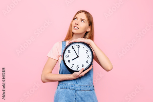 A pensive girl with a wall clock in her hands, gazing thoughtfully into the distance against an isolated pink background