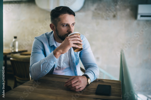 Thoughtful male in casual wear resting at cafeteria on free time drinking hot coffee beverage looking away, serious 20s hipster guy starting day with caffeine holding disposable cardboard cup in cafe