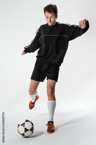 Fototapeta Naklejka Na Ścianę i Meble -  Football player in black sportswear kicking soccer ball to make goal isolated over white background