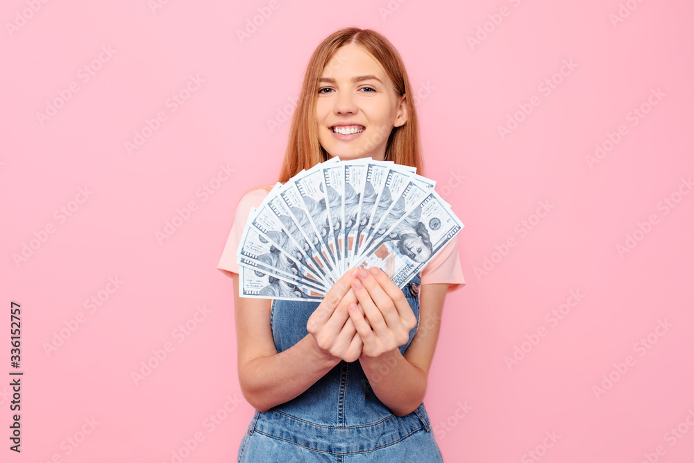 happy young girl holding cash dollar bills on an isolated pink background