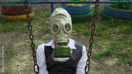 Lonely attractive student guy in suit, in protective rubber green gas mask from virus sit on a rusty old swing on empty playground, looking at the camera on grass background on sunny day close up.  photo