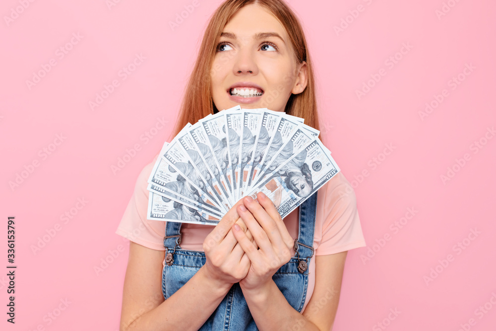 thinking attractive girl holding cash dollar bills, looking away, on a pink background thinking attractive girl holding cash dollar bills, looking away, on a pink background