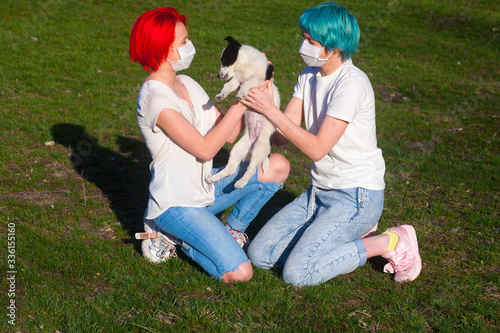 Blue hair young woman and red hair teenage girl play with puppy dog outdoor. Both wear medical mask