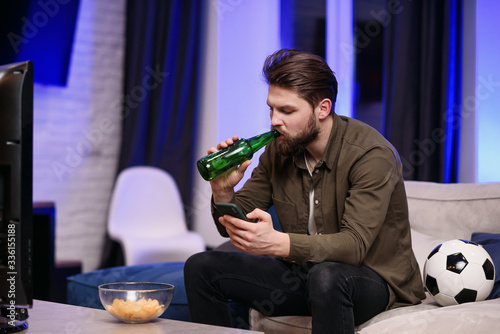 Slow motion of good-looking modern 30-aged guy with beard which sitting in front of tv and periodically watching football match and apps on his mobile while drinking beer photo