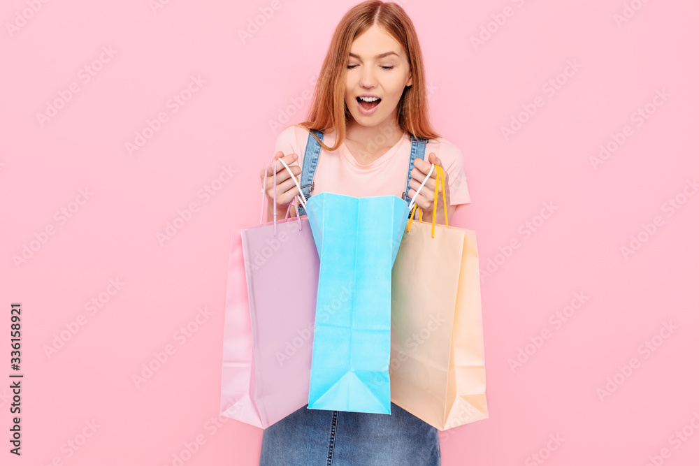 An image of a beautiful shocked surprised young stylish woman posing isolated on a pink background, holding shopping bags