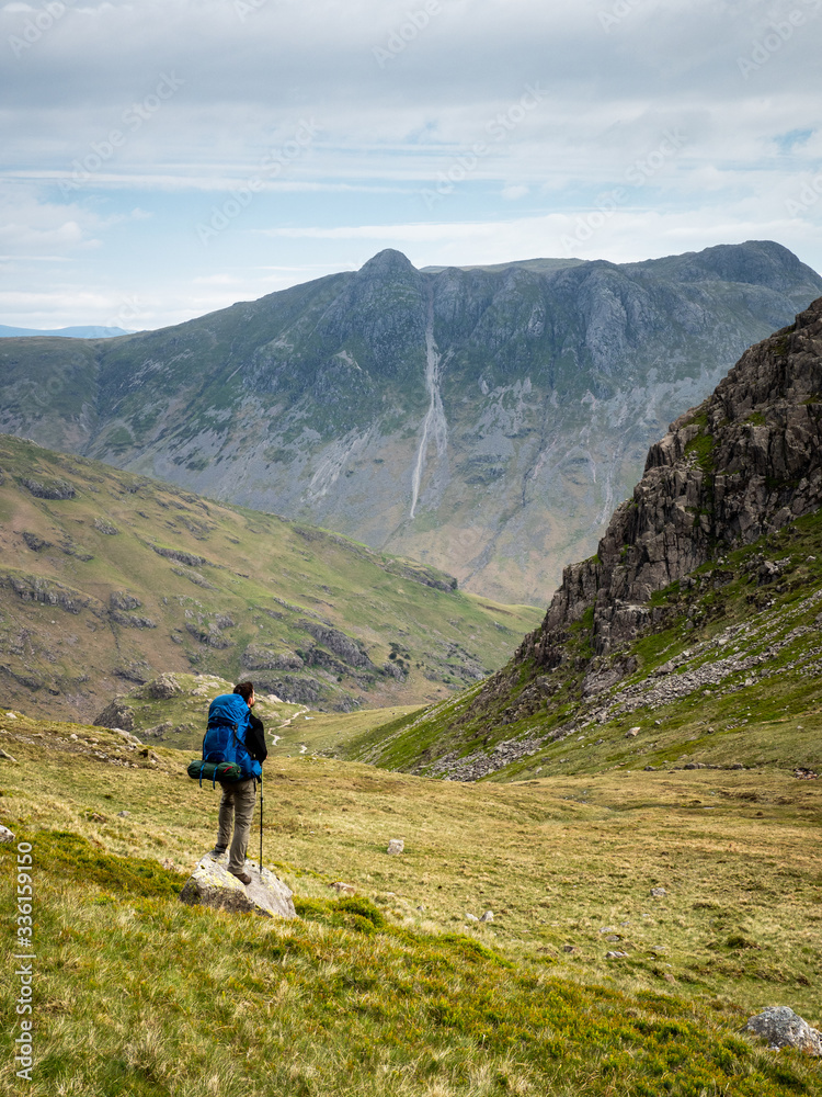 A walker looks towards the Langdale valley and Pike of Stickle beyond Black Wars, cliffs below Pike o' Blisco, a fell in the Lake District, Cumbria, England.