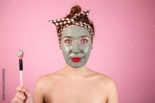 Happy girl with a clay mask on her face on a pink background with a cosmetic brush in her hand