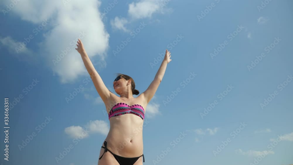 joyful slim girl in bikini sunglasses stands raising arms up enjoying holidays against blue sky with white clouds low angle shot joyful slim girl in bikini sunglasses stands raising arms up enjoying holidays against blue sky with white clouds low angle shot