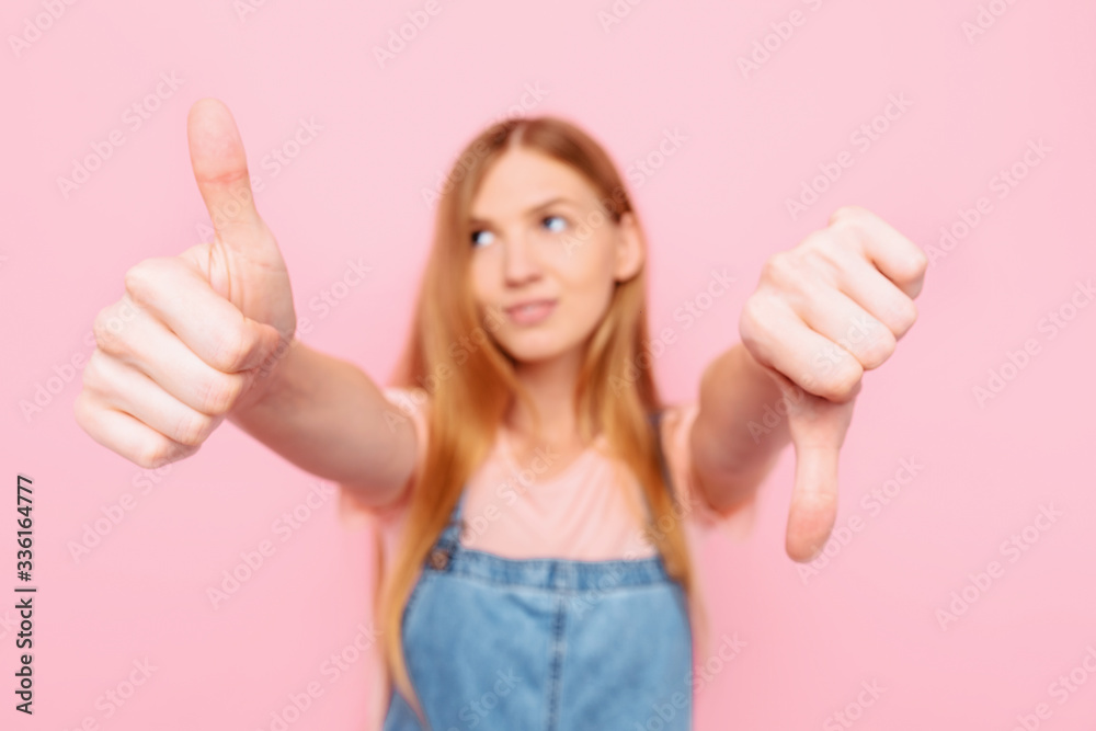 A young attractive woman makes a good-bad sign. Undecided between Yes or no, on an isolated pink background A young attractive woman makes a good-bad sign. Undecided between Yes or no, on an isolated pink background