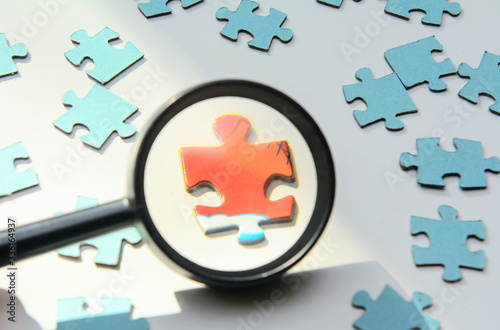 On a white background blue puzzles. A red puzzle is visible through a magnifying glass. Concept - leader selection, team, coronovirus test, patient identification photo