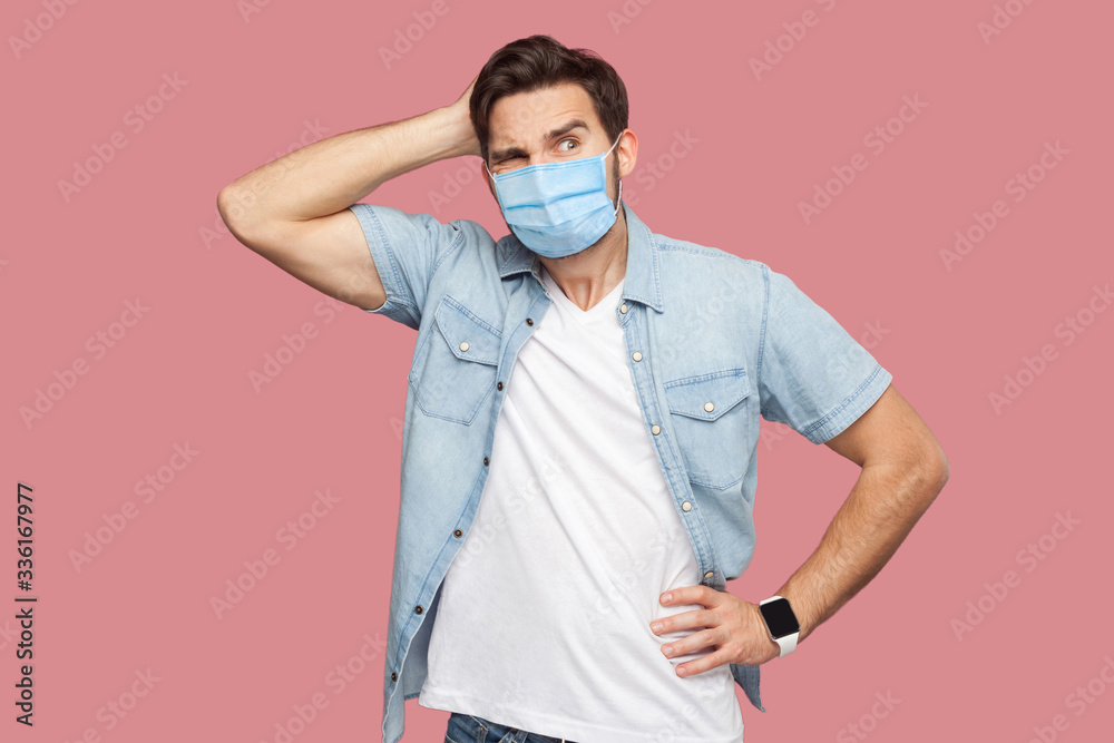 Portrait of young man with surgical medical mask in blue casual style shirt standing, scratching his head, looking away and thinking what to do. indoor studio shot, isolated on pink background. Portrait of young man with surgical medical mask in blue casual style shirt standing, scratching his head, looking away and thinking what to do. indoor studio shot, isolated on pink background.